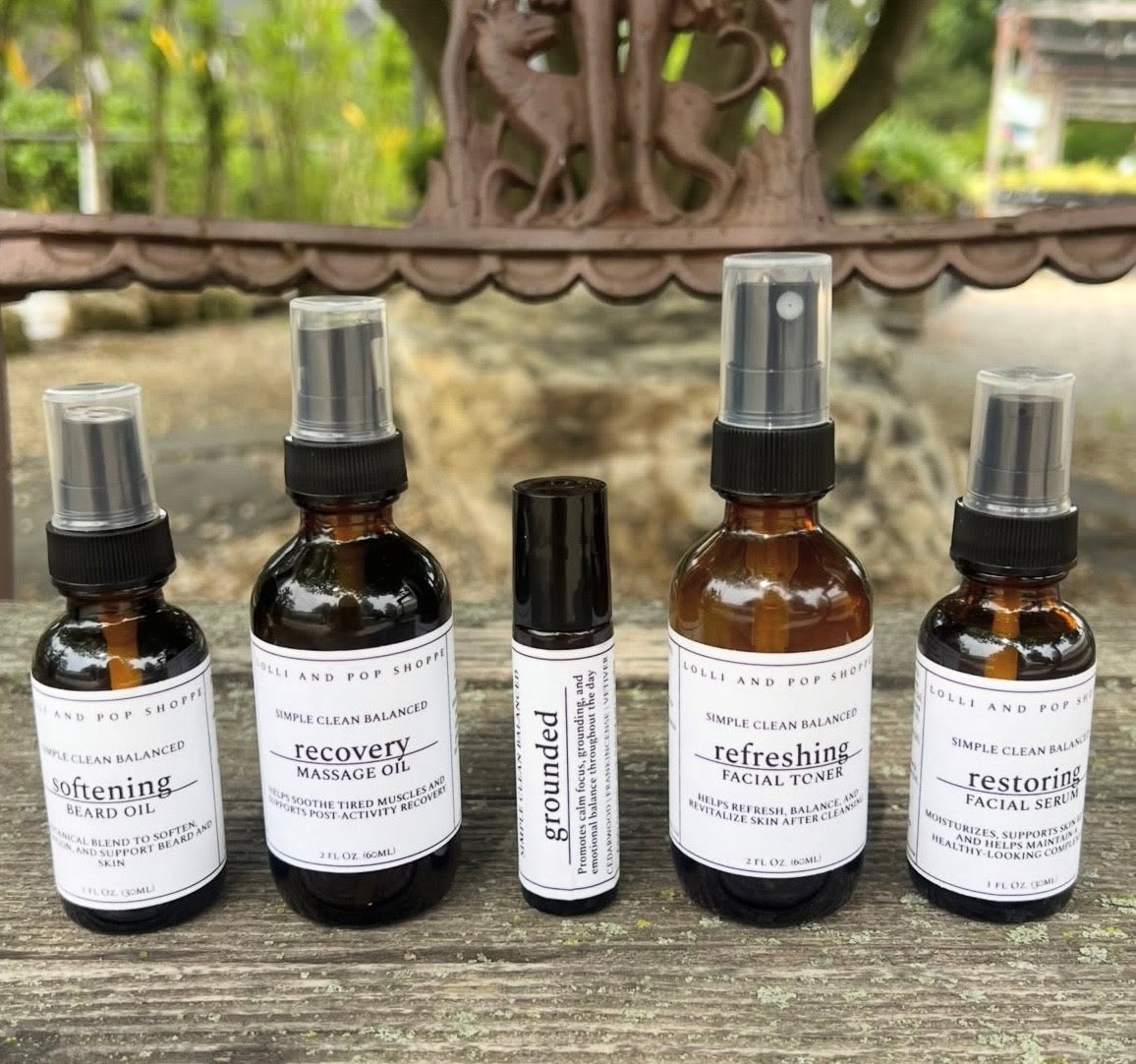Five amber glass skincare and massage oil bottles with white labels are arranged on a weathered wooden surface outdoors. The set includes beard oil, recovery massage oil, a small roller bottle, a facial toner, and a facial serum.