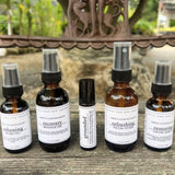 Five amber glass skincare and massage oil bottles with white labels are arranged on a weathered wooden surface outdoors. The set includes beard oil, recovery massage oil, a small roller bottle, a facial toner, and a facial serum.