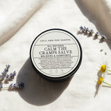 Round container of 'All Natural Calm the Cramps Salve' from Lolli and Pop Shoppe on a wooden surface.