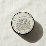 Round container of 'All Natural Calm the Cramps Salve' from Lolli and Pop Shoppe on a wooden surface.