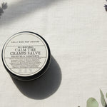 Round container of 'All Natural Calm the Cramps Salve' from Lolli and Pop Shoppe on a wooden surface.