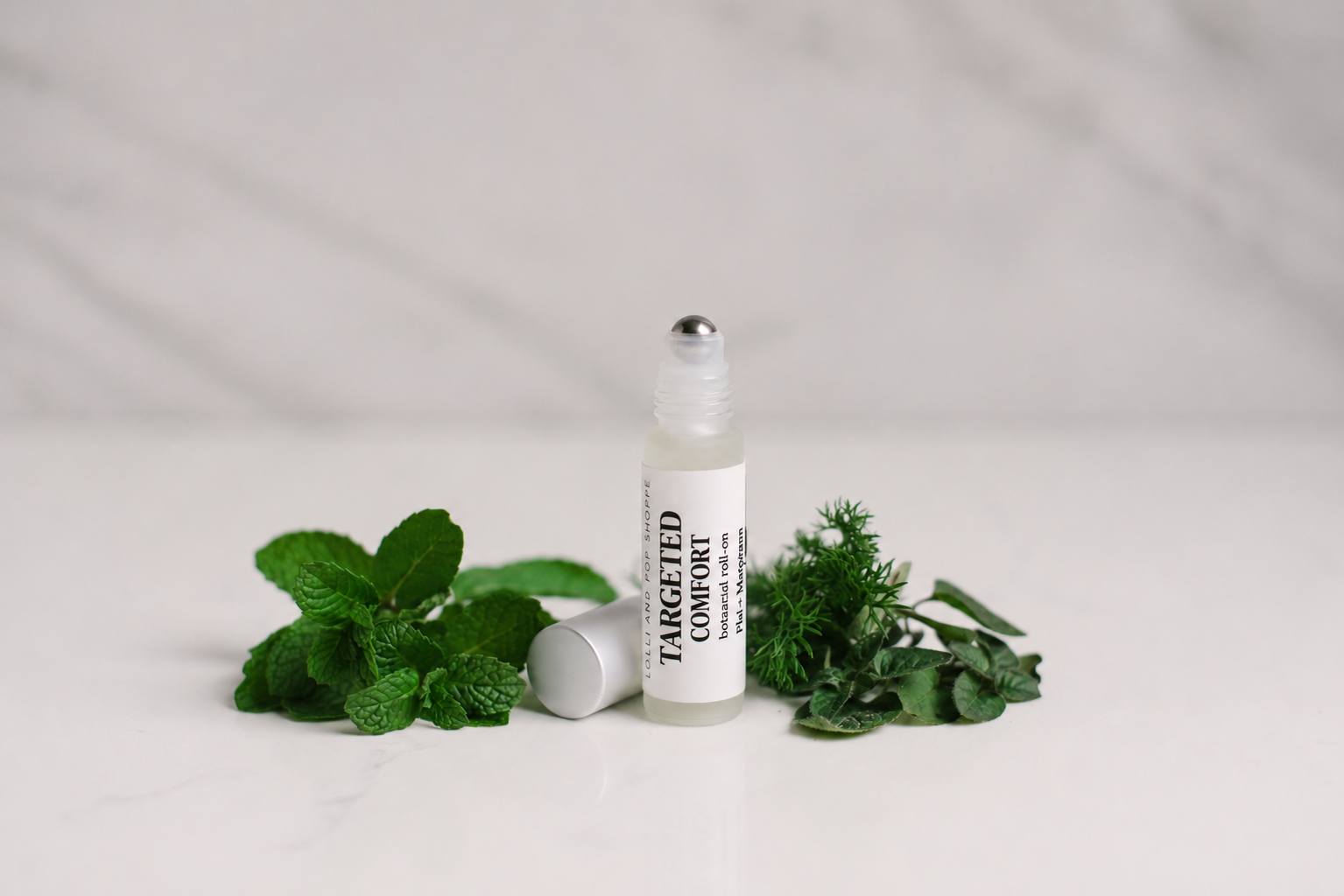 A small clear roll-on bottle labeled TARGETED COMFORT with a white cap, styled on a light marble surface with green mint and herb leaves around it.