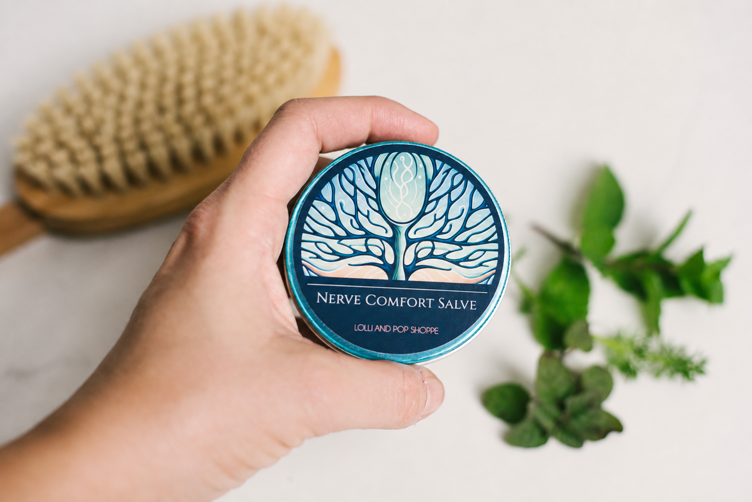 A hand holds a round tin of Nerve Comfort Salve against a light background, with a wooden brush and green leaves blurred in the background.