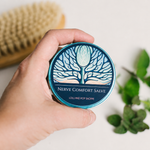 A hand holds a round tin of Nerve Comfort Salve against a light background, with a wooden brush and green leaves blurred in the background.