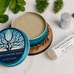 A styled flat-lay of a natural salve product showing an open teal tin of 'Nerve Comfort Salve' with its lid beside it, a silver cap, and a white rollerball bottle labeled 'Targeted Comfort Natural Roll-on' on a white marble surface surrounded by mint leaves and pine sprigs.