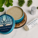 A styled flat-lay of a natural salve product showing an open teal tin of 'Nerve Comfort Salve' with its lid beside it, a silver cap, and a white rollerball bottle labeled 'Targeted Comfort Natural Roll-on' on a white marble surface surrounded by mint leaves and pine sprigs.